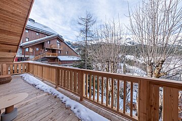 Wooden balcony with patches of snow overlooking bare winter trees, a rustic chalet, and snow-covered mountains.