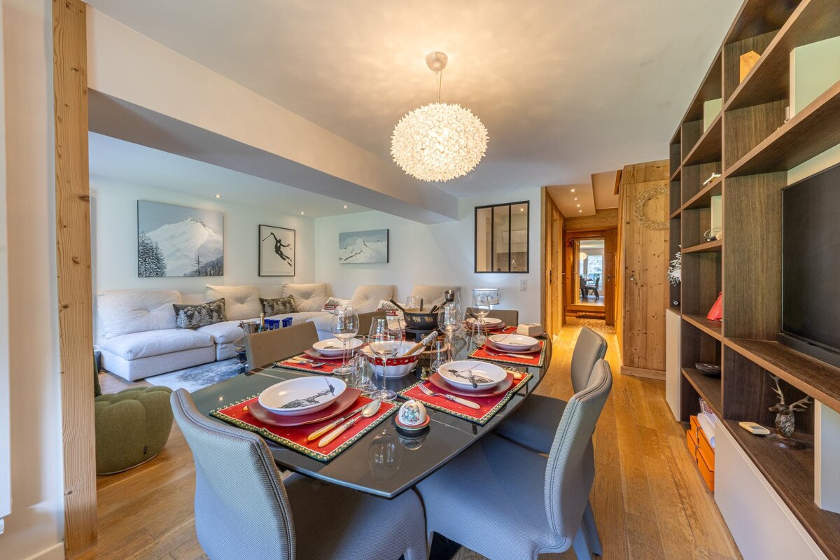A chic chalet-style dining room and living area. A set table with red placemats leads to sofas and mountain art. Warm wood accents complete the cozy, modern space.