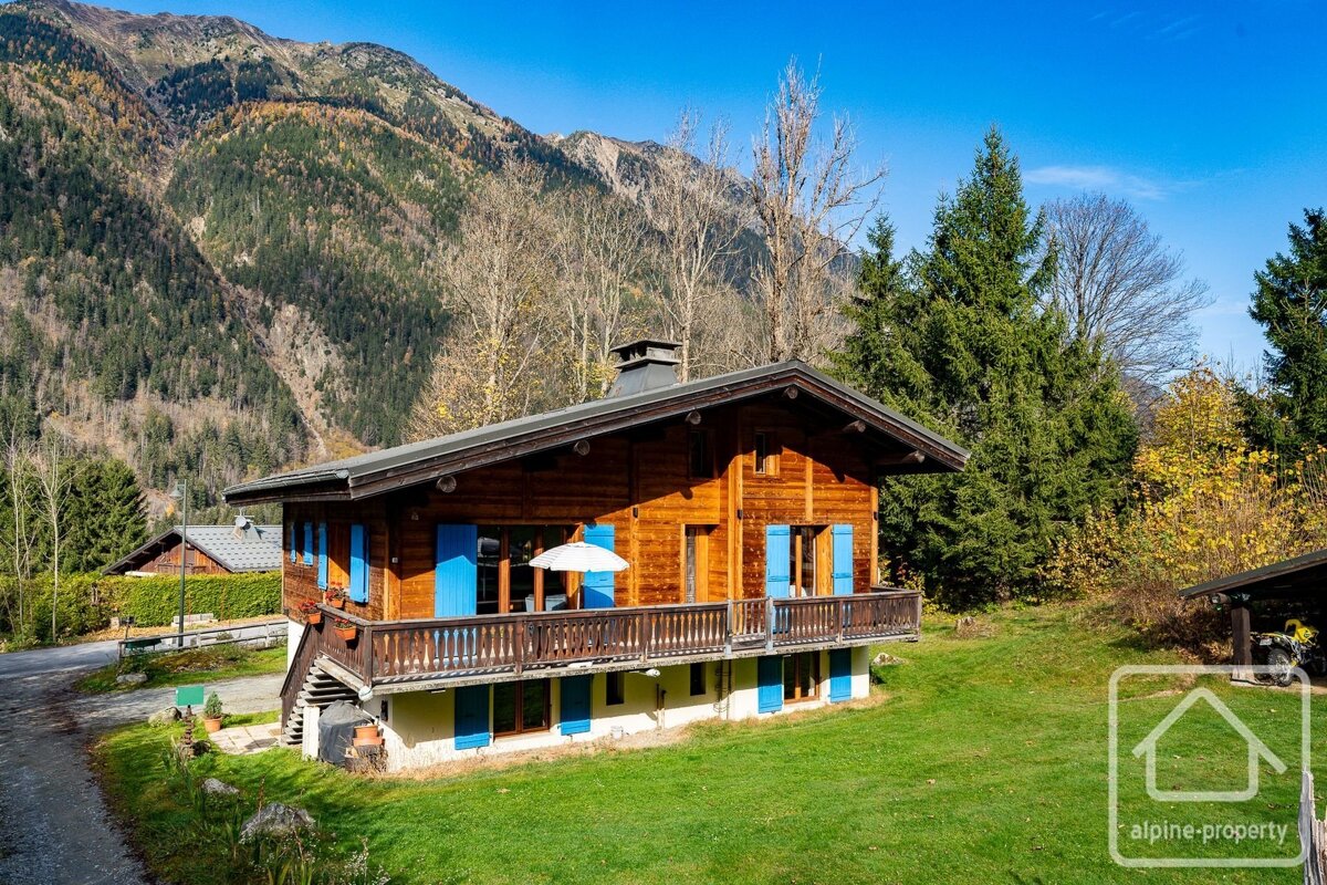 A beautiful wooden chalet with blue shutters and a balcony, nestled on a green lawn with majestic forested mountains in the background under a clear sky.