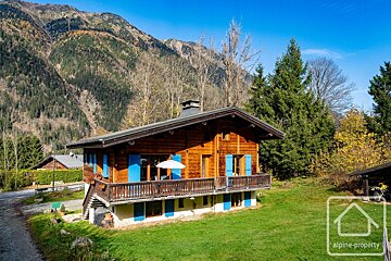 A wooden alpine chalet with blue shutters stands in a grassy clearing, framed by pine trees and rugged mountains.