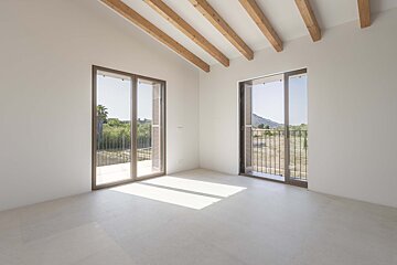 Bright, empty room with white walls, exposed wooden ceiling beams, and two large windows with shutters, offering views of a sunny, green landscape with hills.