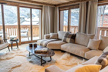 A cozy chalet living room featuring a cream sectional sofa, plush sheepskin rug, and large glass doors revealing a stunning snowy mountain view.