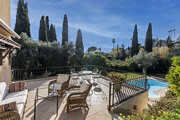 A sunny outdoor patio with wicker seating, dining table, and a pool, surrounded by lush green trees including tall cypress under a bright blue sky.