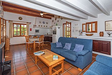 A cozy, open-plan living room and kitchen featuring blue sofas, a dining area, terracotta floor tiles, and rustic wooden details.