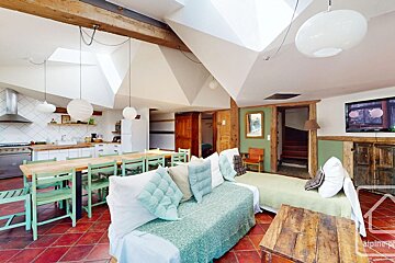 A rustic, open-plan room featuring a kitchen, a long dining table, and a living area, all under sloped ceilings with wood beams and red tiled floors.