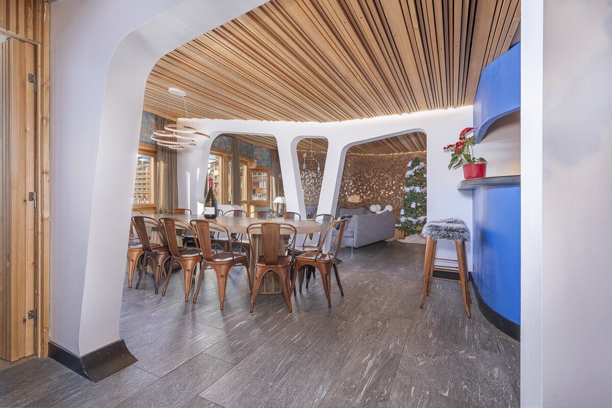 A modern, open-plan room with a dining area, wooden slatted ceiling, and curved white walls leading to a cozy living space with a Christmas tree and a blue bar.