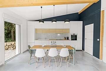 Sleek modern kitchen/dining room with white cabinets, wood accents, dark blue wall, and large sliding door to a green outdoor space.
