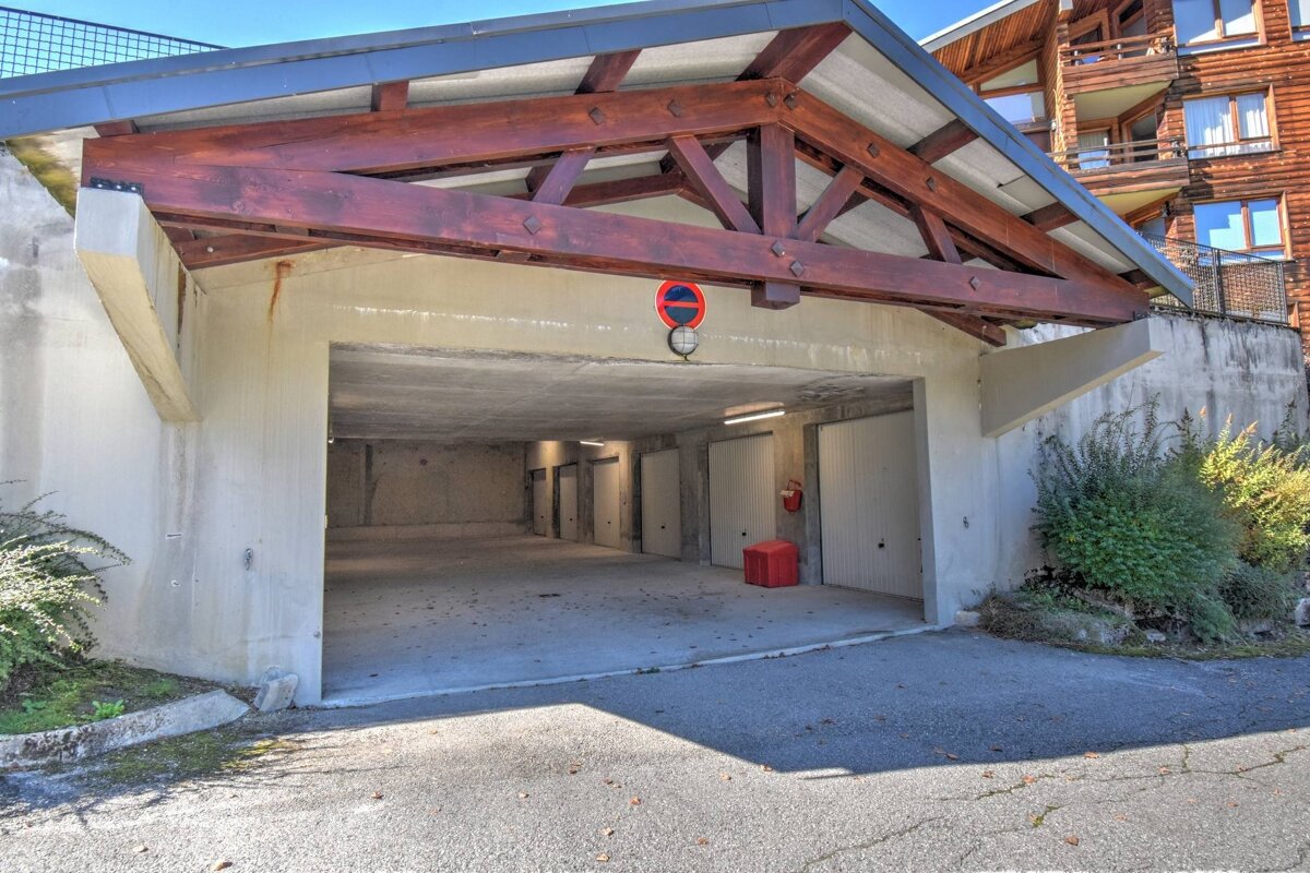 A covered parking garage entrance with a prominent wooden beam roof, revealing individual garage doors inside. An asphalt driveway leads to it, with foliage nearby.