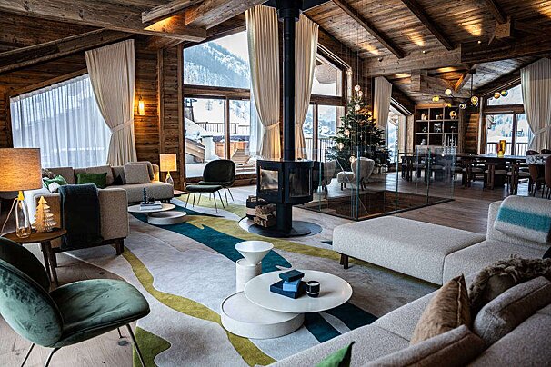 A cozy, luxurious chalet living room with wooden interior, modern fireplace, Christmas tree, stylish furniture, and large windows revealing snowy mountains.