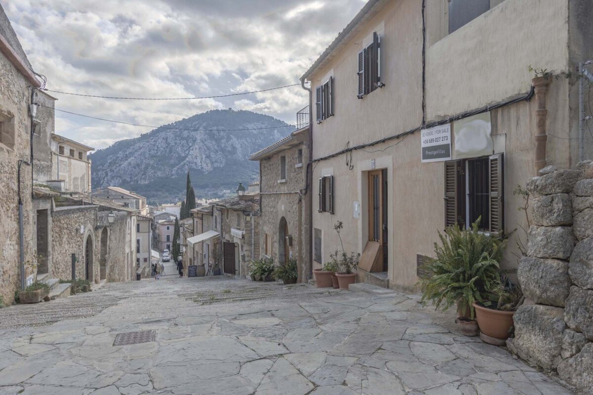 A picturesque stone-paved street in an old European village, with traditional buildings, mountains in the distance, and a cloudy sky.