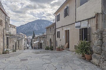 A picturesque stone-paved street in an old European village, with traditional buildings, mountains in the distance, and a cloudy sky.