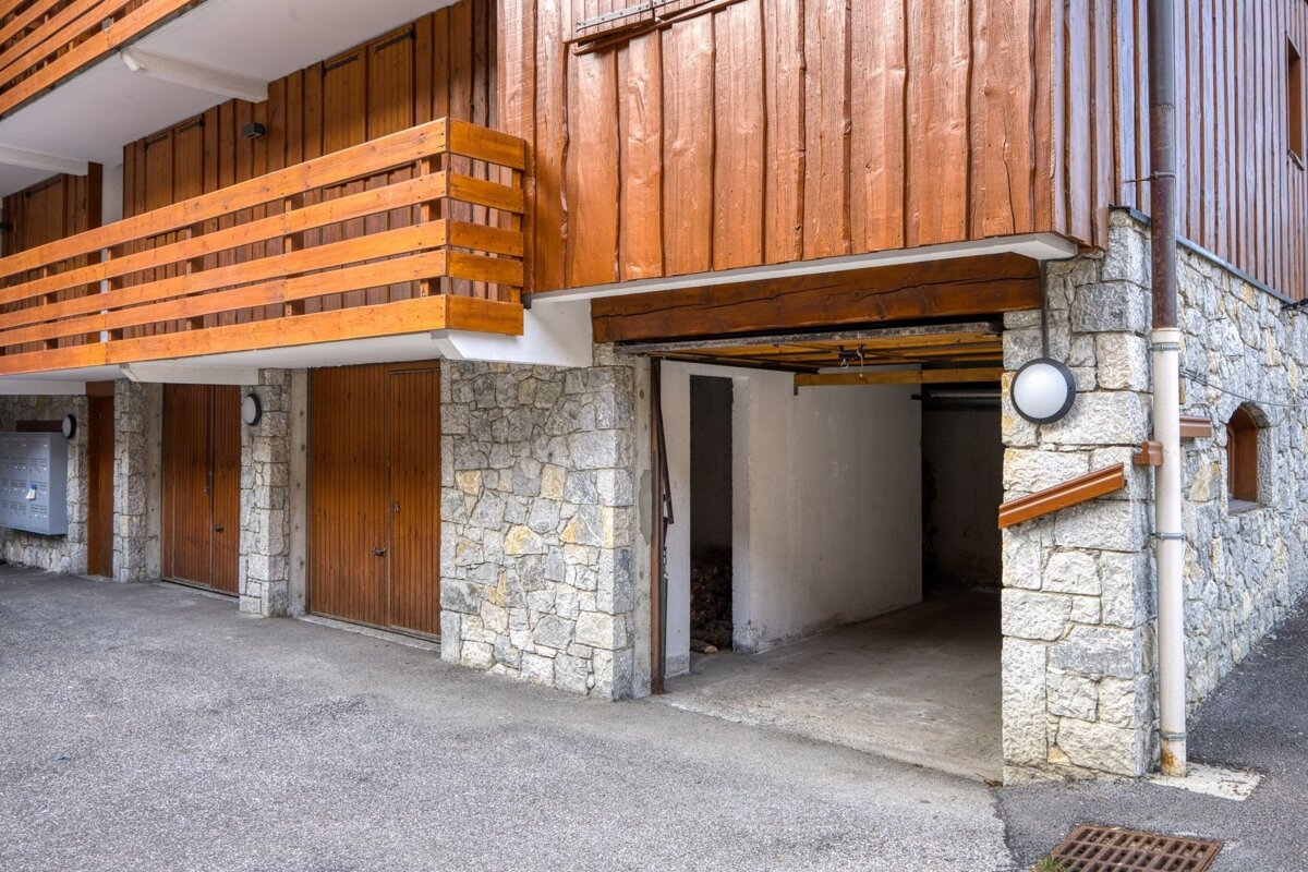 A building with a stone foundation and wooden upper stories features multiple garage doors (one open) and a wooden balcony overlooking a paved area.