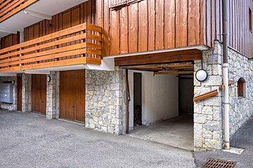 A building with a stone foundation and wooden upper stories features multiple garage doors (one open) and a wooden balcony overlooking a paved area.