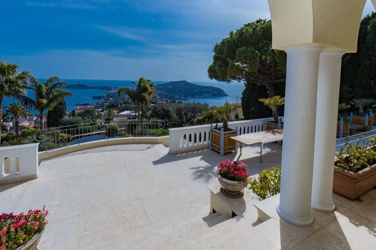 Elegant patio with panoramic sea and coastal views, featuring white balustrades, columns, lush palms, and outdoor seating under a clear blue sky.