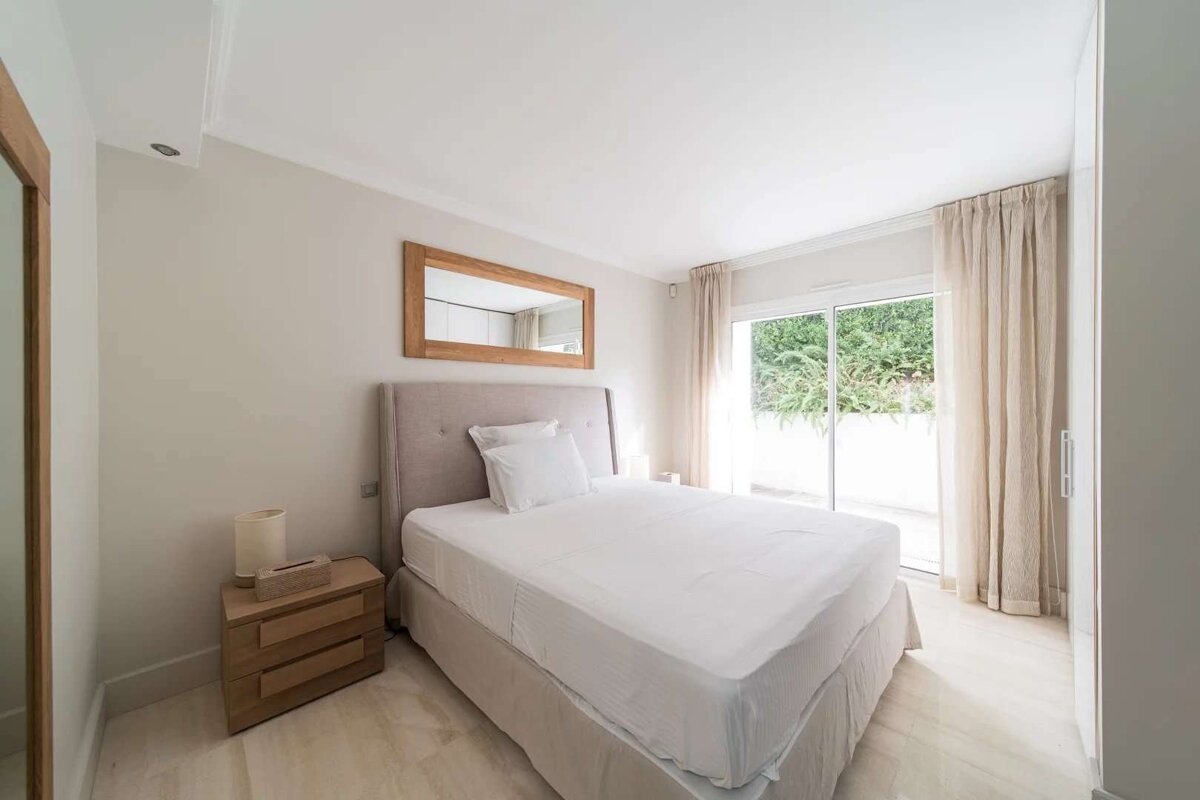 A bright, neutral-toned bedroom with a large bed, wooden mirror, and sliding glass doors opening to greenery.