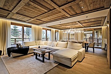 Luxurious living/dining room with a large sectional sofa, rustic wooden coffered ceiling, hardwood floors, large windows, and a grand chandelier.