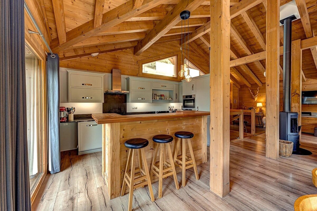 Cozy, modern wooden chalet interior with a kitchen, bar, and wood stove, featuring prominent exposed timber beams and light gray cabinets.