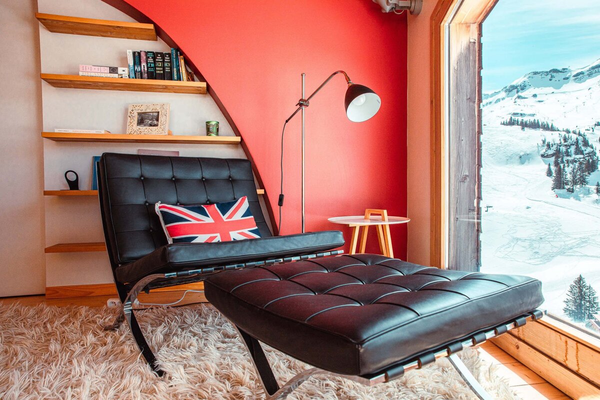 A modern interior with a black leather chair, Union Jack cushion, and red wall, overlooking a snow-covered mountain vista through a large window.