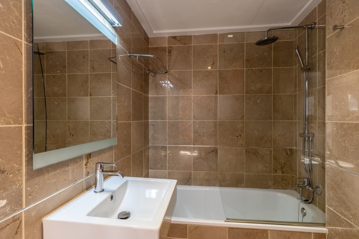 A bathroom with brown tiled walls, a white sink, and a white bathtub with a shower and glass screen. A mirror is above the sink.