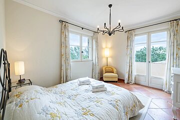 A bright bedroom with a floral bed, terracotta floors, large windows with outdoor views, a yellow armchair, and an elegant black chandelier.