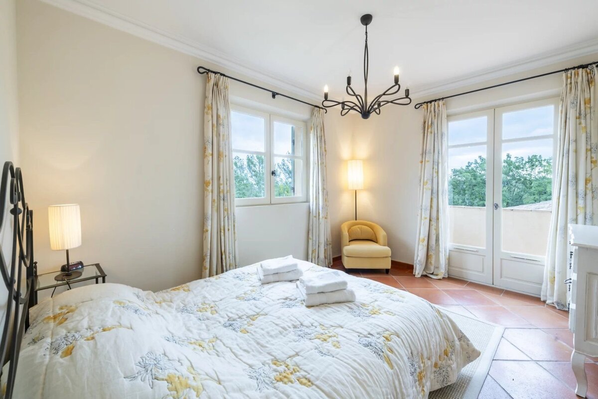 A bright bedroom with a queen bed, a yellow armchair, a black chandelier, and glass doors opening to a view outside.