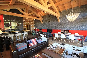 Rustic-modern open-plan chalet with wooden beams, stone wall, red fireplace, kitchen, dining area, and living room with leather sofas.