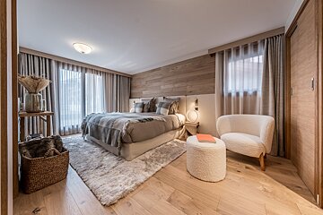 A cozy, modern bedroom with a large bed, wood accent wall, neutral tones, plush rug, and comfortable seating by a window with sheer curtains.