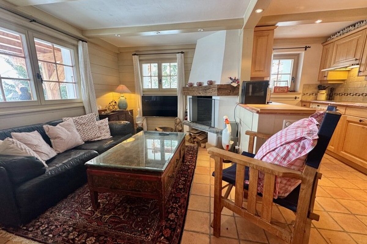 A cozy, rustic living room featuring a black leather sofa, a glass coffee table, a fireplace, and an adjoining kitchen area.