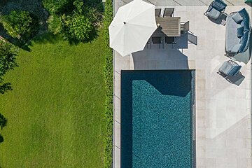 Aerial view of a backyard featuring a swimming pool, a patio with an umbrella and outdoor furniture, and a vibrant green lawn with hedges.