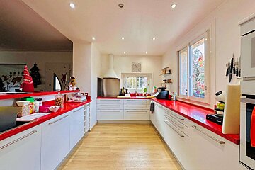 A bright, modern kitchen with white cabinets and bold red countertops. Features light wood floors and festive holiday decorations.
