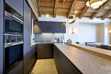 A modern kitchen featuring dark cabinetry, stone countertops, and a rustic wood-beamed ceiling, opening to an elegant dining area.