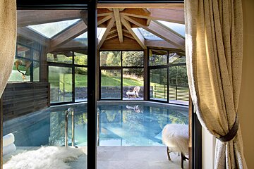 A stunning indoor swimming pool with panoramic glass walls and a wooden beamed ceiling, opening to a lush green outdoor landscape.