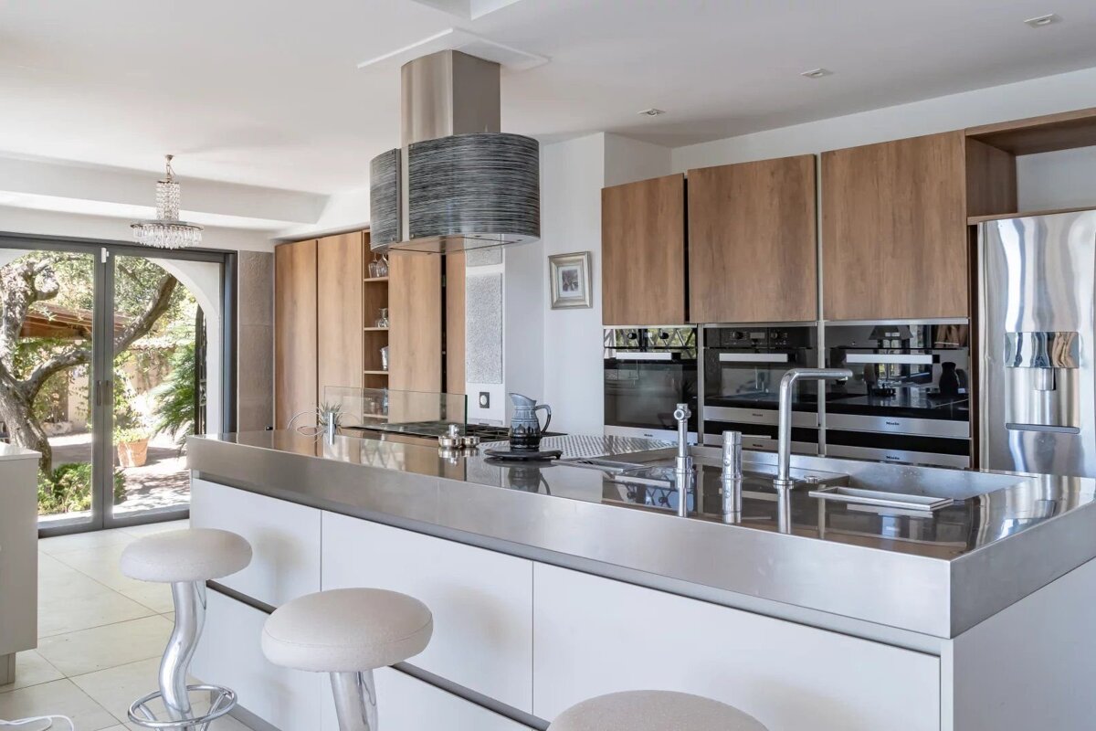 Modern kitchen with a large stainless steel island, white bar stools, wooden cabinets, built-in ovens, and a unique hanging range hood.