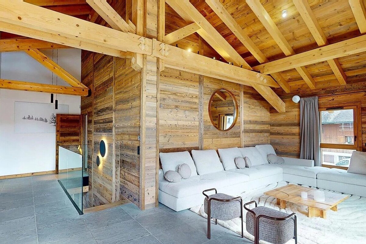 Cozy wooden chalet living room with vaulted timber ceilings, a white sectional sofa, modern stools, and a glass-railed staircase.