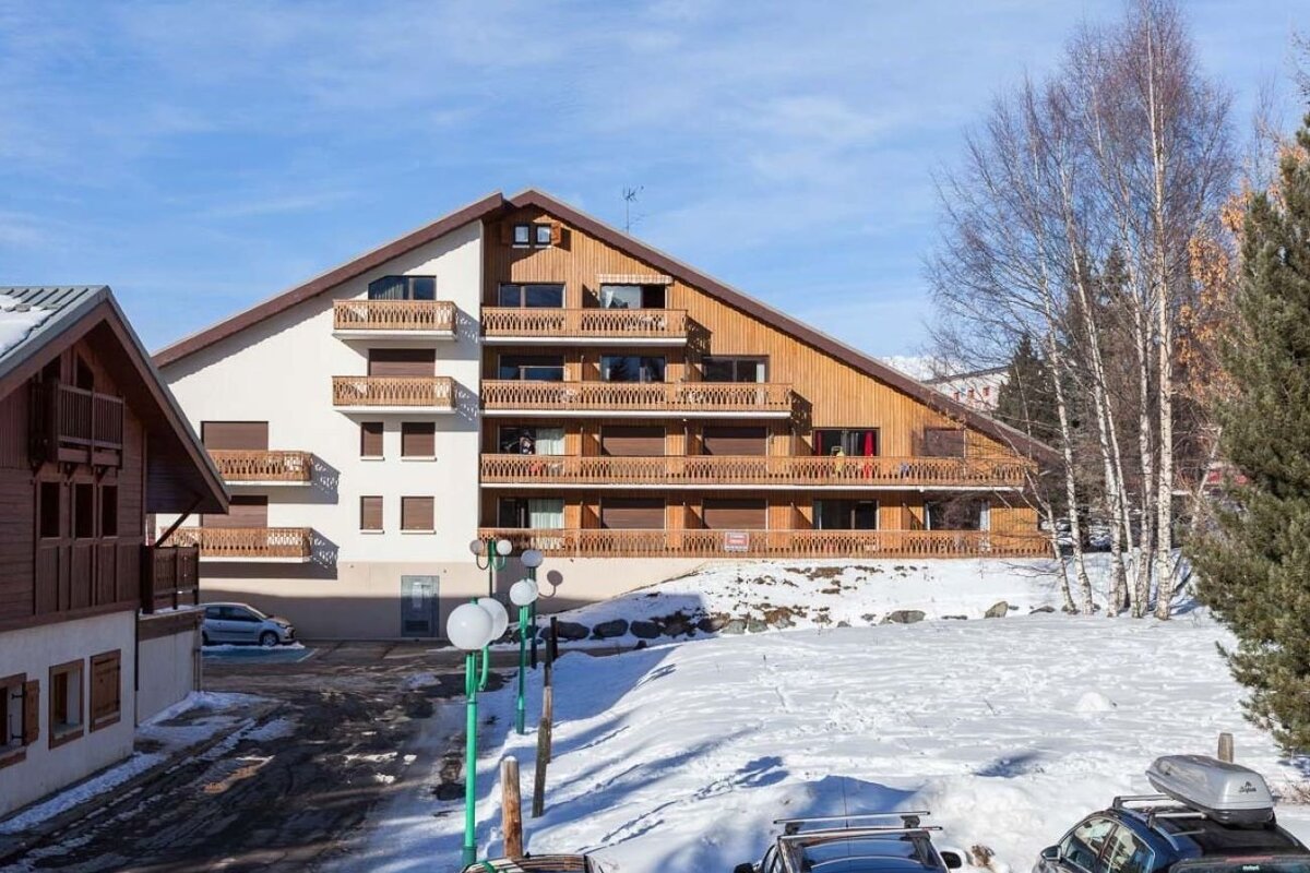 A large alpine chalet building with wooden balconies set in a snowy landscape with bare trees and a clear blue sky.