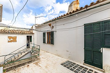Outdoor courtyard with white walls, green shutters, and stairs. Overhead wires, tiled roofs, and a cloudy sky complete the rustic scene.