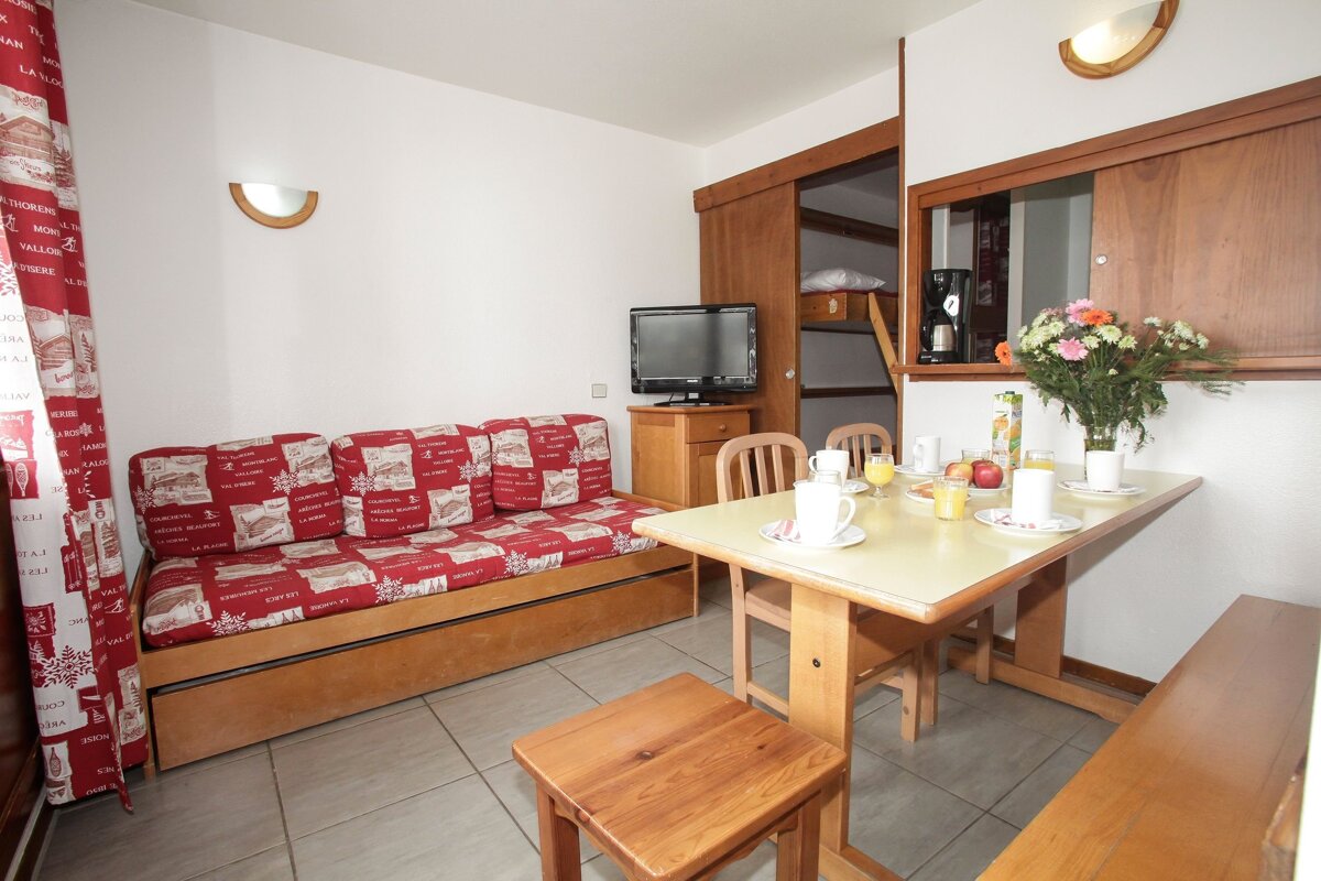 An alpine-style studio featuring a red sofa bed, dining table with breakfast, TV, and bunk beds visible in an open alcove.