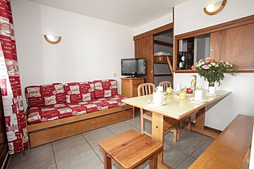 An alpine-style studio featuring a red sofa bed, dining table with breakfast, TV, and bunk beds visible in an open alcove.