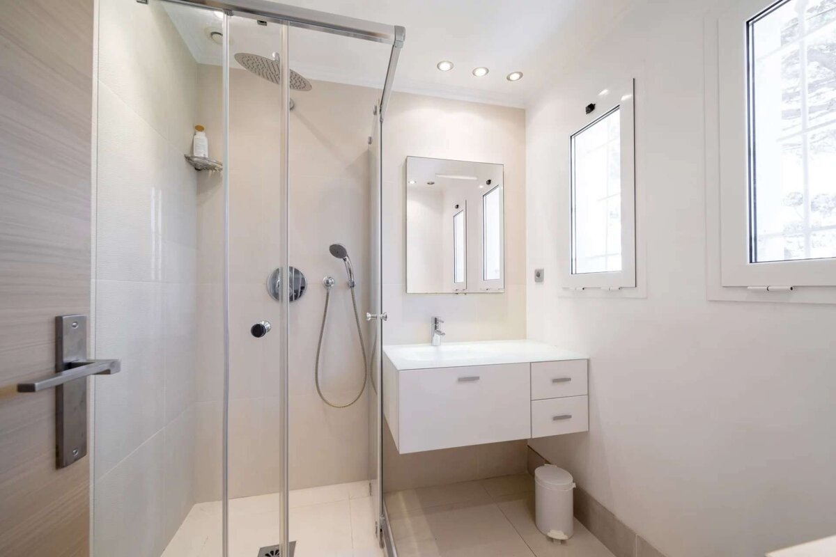 Modern, minimalist bathroom with a glass shower, floating vanity, mirror, and window, featuring clean white and beige tones.