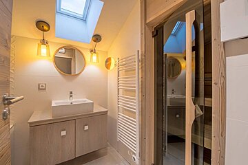 Warm, modern bathroom with a wooden vanity, skylight, round mirror, and an adjacent home sauna with a glass door.