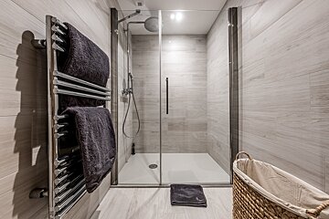 A bright, modern bathroom featuring a walk-in glass shower, a chrome heated towel rail with dark towels, and light grey wood-look tiled walls.