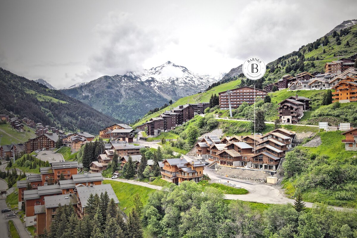 An alpine village with numerous wooden chalets dotting lush green mountainsides, under cloudy skies and snow-capped peaks. A Barnes logo is visible.