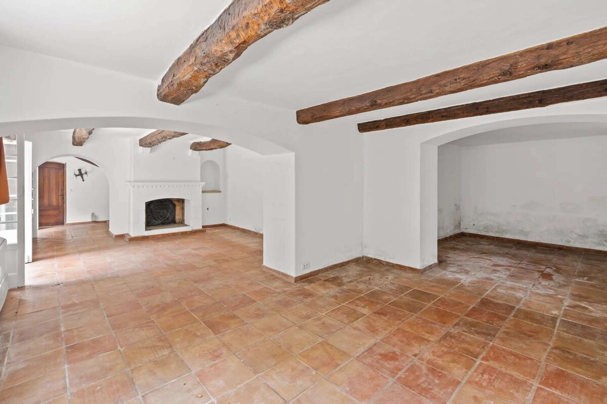 Empty room with white walls, terracotta floor, and rustic wooden ceiling beams. Features a fireplace and arched entryways, with some dampness visible.