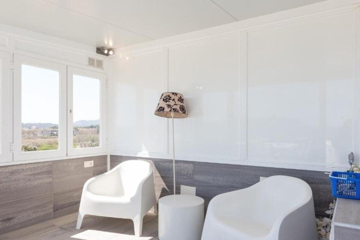A bright, minimalist sunroom with two white armchairs, a small round table, and a floral floor lamp.