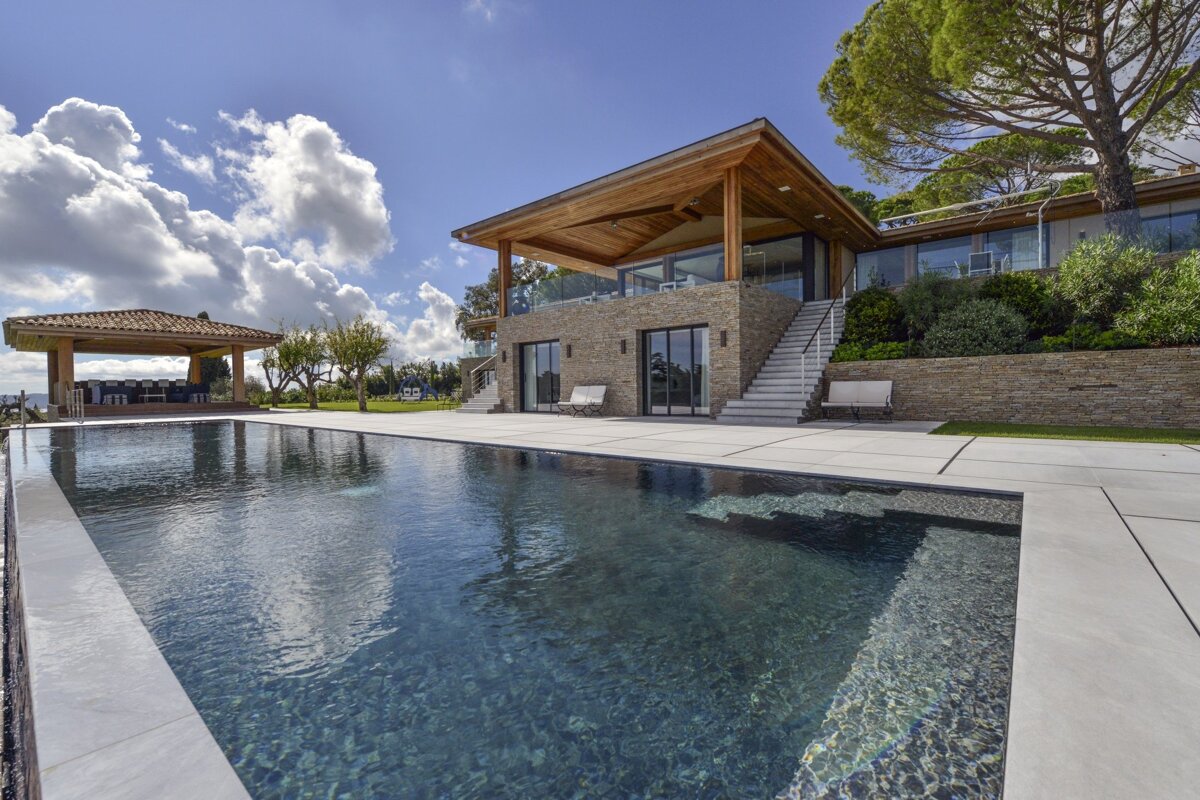 A stunning modern villa with stone walls and wooden accents overlooks a tranquil, dark-tiled swimming pool. Lush landscaping and a bright, cloudy sky complete the scene.