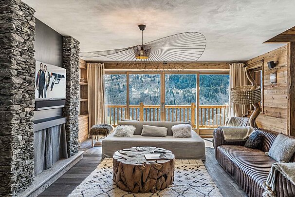 A luxurious mountain living room with rustic stone and wood decor, a unique ceiling light, and plush seating. Large windows offer stunning views of snow-covered mountains.