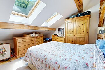 Cozy attic bedroom with slanted ceiling, two skylights, exposed wooden beams, wooden dressers, a wardrobe, and a bed with a patterned duvet.