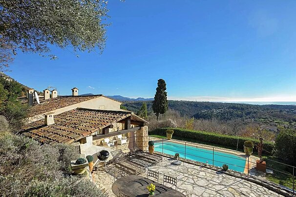 Sunny Mediterranean villa with a pool and stone patio, offering panoramic views of green hills and the distant sea under a clear blue sky.