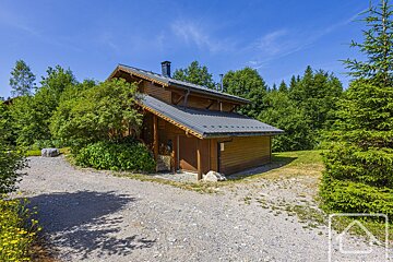 A wooden chalet with a gray roof stands beside a gravel path, surrounded by lush green trees and bushes under a clear blue sky on a sunny day.