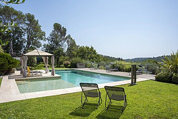 An outdoor swimming pool surrounded by a green lawn, lounge chairs, a gazebo, and lush trees under a clear blue sky.